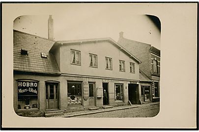 Hobro, bygning med Hobro Mejeri-Udsalg, Jens Fæsters Tobak og Cigarhandel, samt L. C. Lohmann's Pakfarveforretning. Fotokort u/no.